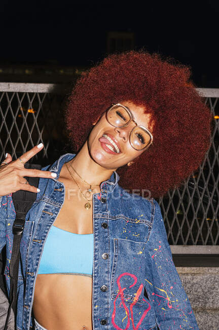 Mujer positiva con peinado afro mostrando gesto de paz mientras mira a la cámara con la lengua cerca de la valla en el tiempo de la noche - foto de stock