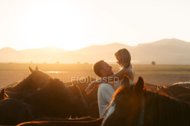 Side view of happy female admiring sunset over mountains while being raised by loving man among calm horses in Turkey field — Stock Photo