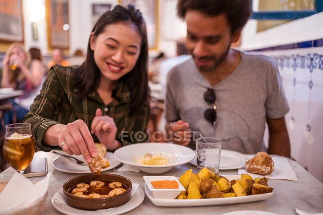 Content young Asian female millennial dipping bread into sauce of appetizing gambas al ajillo dish with shrimps while having dinner with positive ethnic boyfriend — Stock Photo