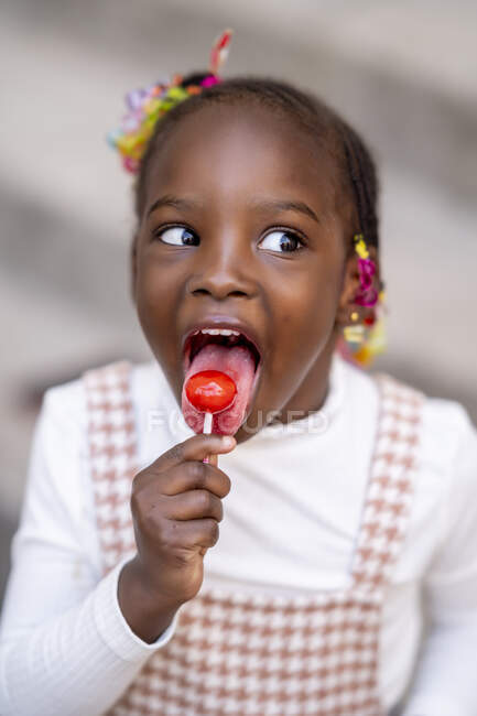 De cima de menina afro-americana feliz na roupa da moda em pé com a boca aberta e lambendo pirulito doce contra fundo borrado — Fotografia de Stock