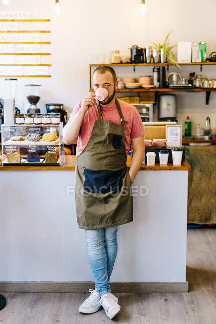 Voller Körper eines ernsthaften männlichen Baristas in Schürze, der in die Kamera schaut, während er heißen Kaffee in der Nähe der Theke in einem modernen Kaffeehaus trinkt — Stockfoto