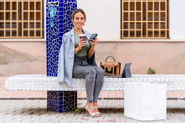 Full body of positive female in stylish outfit text messaging on cellphone and looking at camera while sitting with takeaway coffee on bench with purse and tablet — Stock Photo