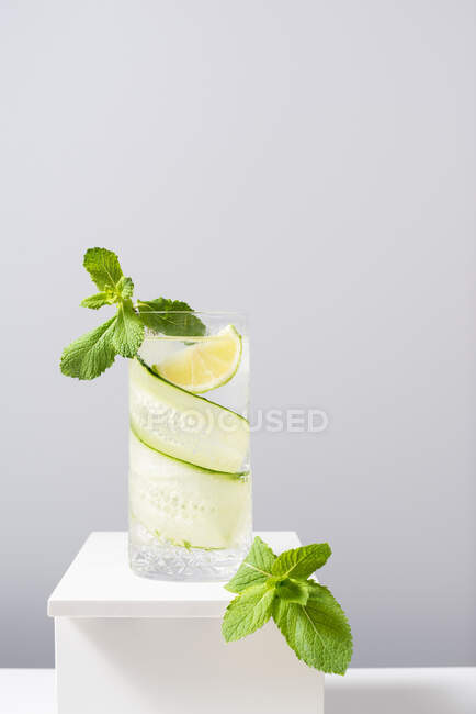 Glass of refreshing gin and tonic with cucumber and lime decorated with mint leaves placed on white table against gray background — Stock Photo