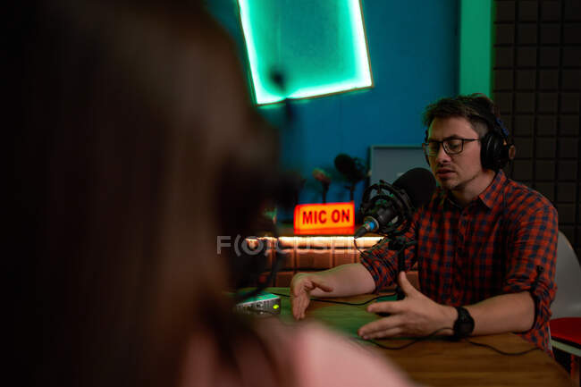 Young male radio host in checkered shirt and headphones sitting at table with microphone and communicating with crop anonymous colleague during podcast recording in studio — Stock Photo