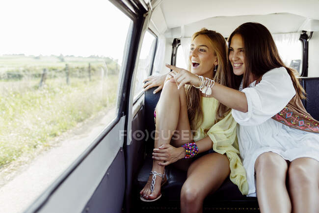 Dos hermosas chicas sonrientes sentadas dentro de una furgoneta vestidas con ropa de verano chicas señalando desde la ventana de una furgoneta - foto de stock