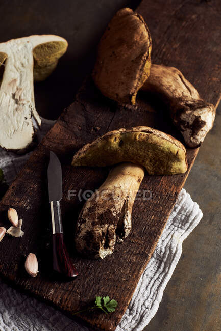 Top view of raw cut Boletus edulis mushrooms on wooden chopping board with garlic and parsley in light kitchen during cooking process — Stock Photo