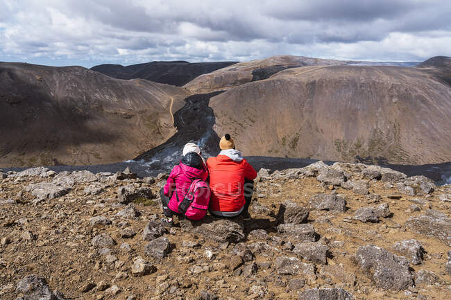 Погляд на нерозпізнану пару в теплому верхньому одязі, що сидить на скелястій землі і захоплюється мальовничими краєвидами активного вулкана Фаградальсфьялл в Ісландії в денний час. — стокове фото