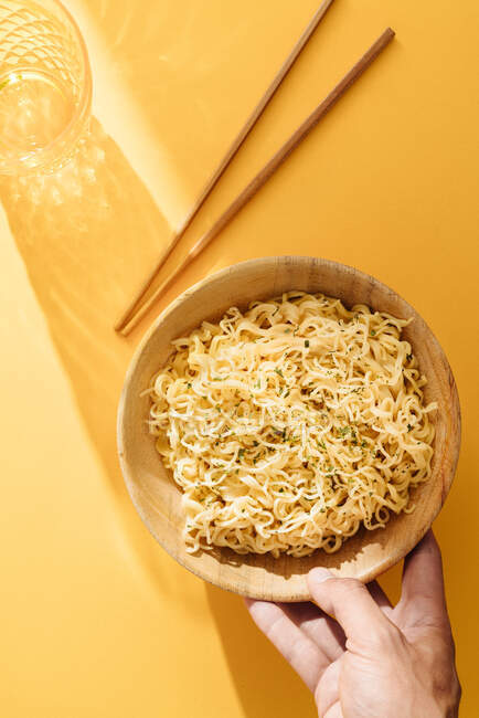 Top view of crop anonymous person holding fresh noodles in bowl placed on yellow background with glass and wooden chopsticks — Stock Photo