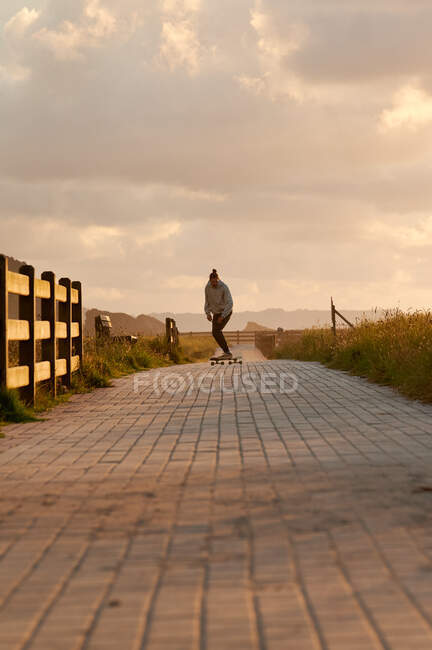 Corpo completo di longboard attivo maschile lungo un lungo sentiero asfaltato sotto il cielo nuvoloso al tramonto della sera — Foto stock