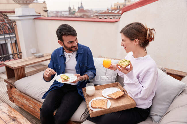 Positive Frau sitzt auf der Couch und isst leckeres Sandwich mit Avocadoscheiben und trinkt ein Glas Orangensaft — Stockfoto