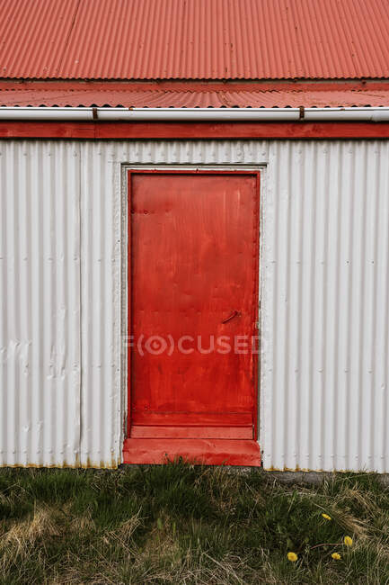 Façade de maison en métal vieilli avec porte rouge et toit sur prairie herbeuse dans la campagne — Photo de stock
