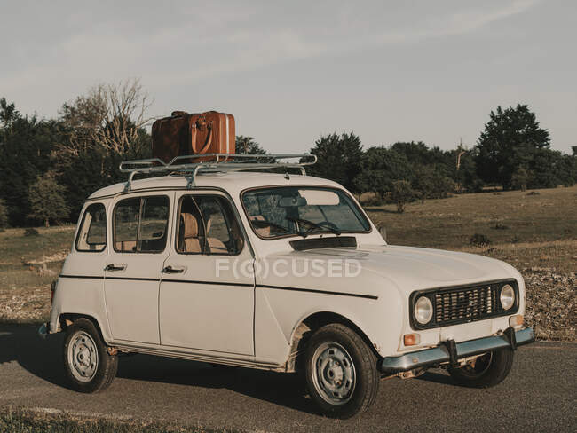 Old timer white automobile with vintage suitcase on roof parked on road near grassy field in nature on summer day — Stock Photo