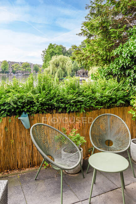 Picturesque view of empty terrace with chairs and small green table fenced off with enclosure made of natural materials with blooming bushes and lake on background in summer on sunny day — Stock Photo