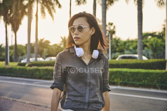 Серйозна азіатка з сучасними білими навушниками, що дивляться у далечінь, стоячи біля дороги на вулиці з зеленими деревами. — стокове фото