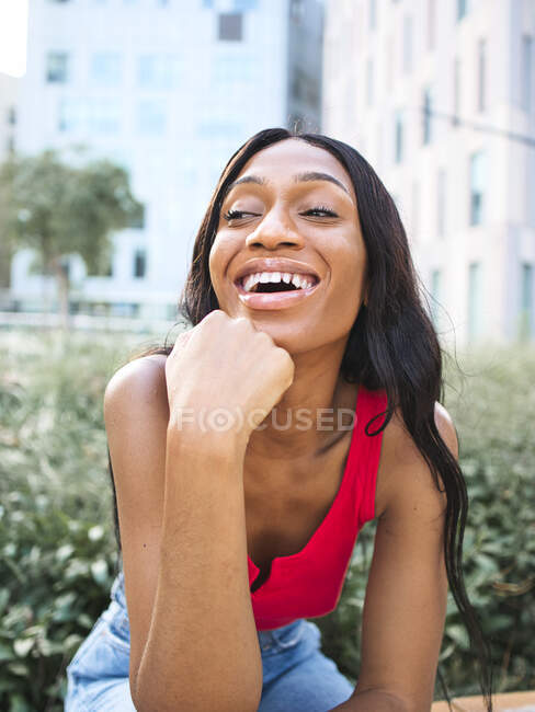 Mujer afroamericana feliz con ropa casual apoyada en la mano con sonrisa y mirando hacia otro lado mientras está sentada en la calle cerca de arbustos verdes - foto de stock