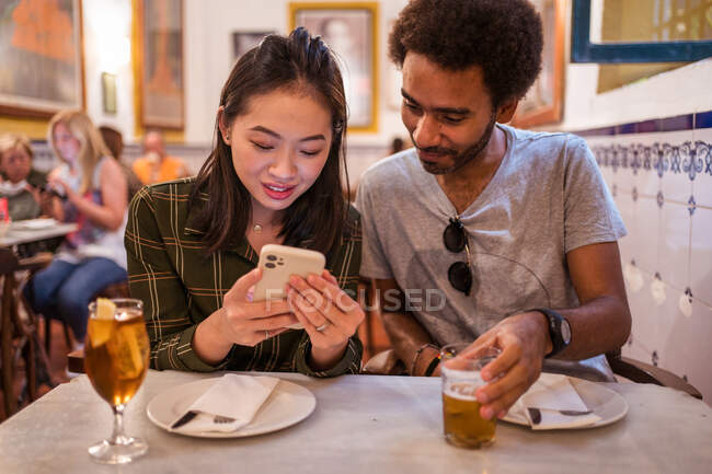 Joven mujer asiática mostrando fotos en smartphone a novio étnico sentado en la mesa en el restaurante - foto de stock