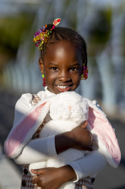 Cheerful African American girl with hairstyle standing and hugging teddy rabbit on street in sunny day — Stock Photo