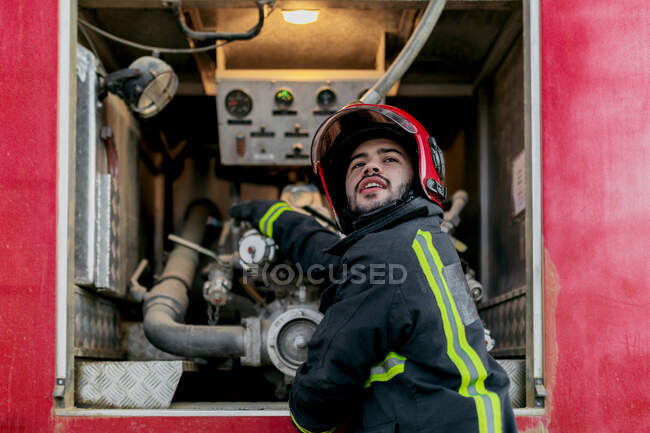 Travailleur portant un uniforme de protection et un casque rouge fonctionnant avec un pipeline situé à l'intérieur du camion de pompiers — Photo de stock