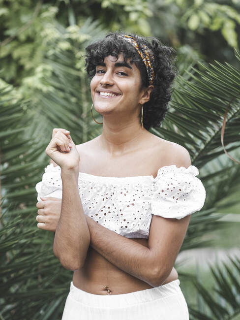Весела чарівна етнічна жінка в пов'язці на кучеряве волосся дивиться на камеру в зеленому літньому саду — стокове фото