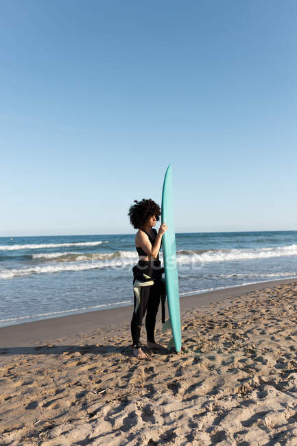 Vista lateral de la joven surfista en traje de neopreno con tabla de surf de pie mirando hacia otro lado en la orilla del mar bañada por el mar ondulante - foto de stock