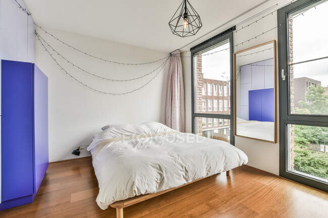 Удобная кровать с белой постельным бельем помещается у стены с висящей гирляндой в стильной спальне со стеклянными дверями в современной квартире — стоковое фото
