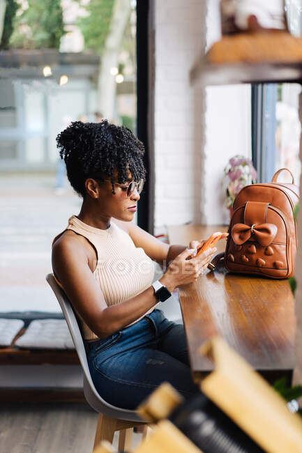 Side view of confident young African American female millennial with dark curly hair in trendy outfit and sunglasses sitting at table in modern cafe and messaging on smartphone — Stock Photo