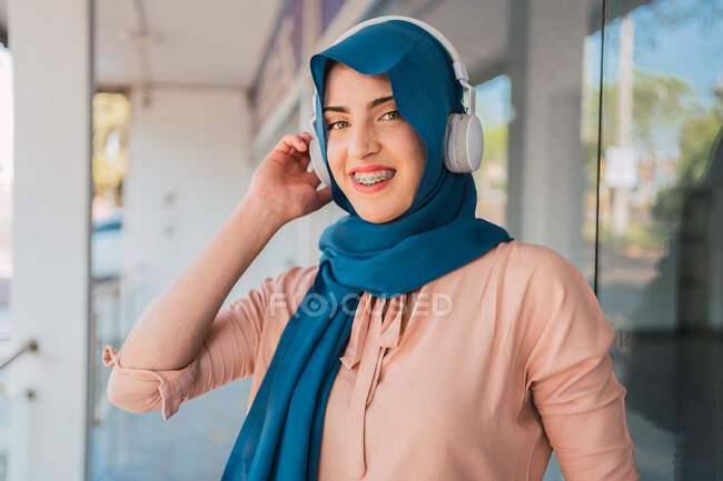 Позитивная арабская женщина в головном уборе и наушниках слушает музыку, смотрит в камеру и наслаждается песнями, стоя на городской улице. — стоковое фото