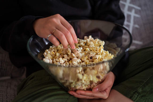 Ritagliato femmina irriconoscibile indossa abiti casual seduto con le gambe incrociate sul divano e mangiare popcorn a casa — Foto stock