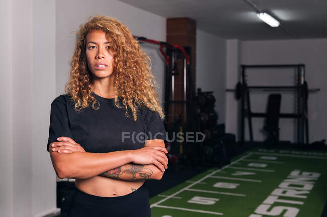 Confident hispanic sportswoman with long curly hair in activewear standing with crossed arms in gym and looking at camera — Stock Photo