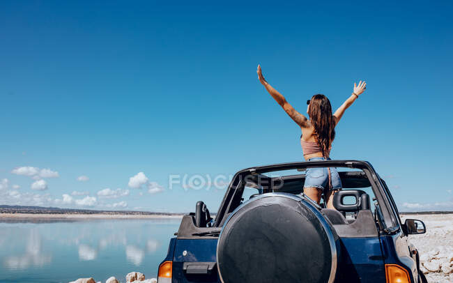 Back view of unrecognizable young tattooed female traveler in top and denim shorts standing with arms raised in convertible SUV car parked on sandy beach and admiring picturesque seascape under cloudless blue sky — Stock Photo