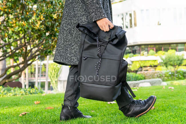 Vista lateral da cultura anônimo empresário masculino em botas e casaco com mochila passeando no prado no parque da cidade — Fotografia de Stock