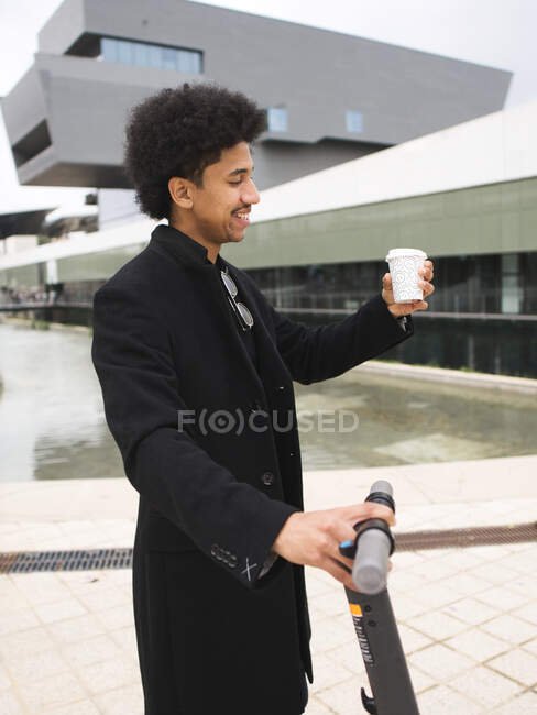 Joven y elegante hombre afroamericano con cabello oscuro rizado en elegante abrigo de pie en la calle con scooter eléctrico y una taza de café para llevar y mirando hacia otro lado - foto de stock