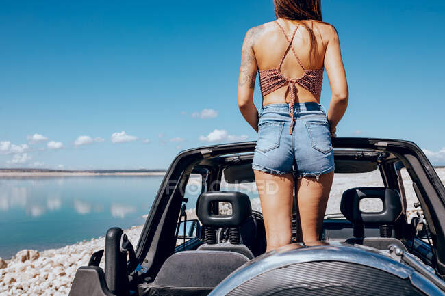 Back view of unrecognizable young tattooed female traveler in top and denim shorts standing in convertible SUV car parked on sandy beach and admiring picturesque seascape under cloudless blue sky — Stock Photo