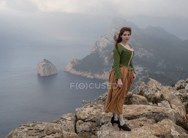 Вид збоку тіла мрійливої жінки в старомодному одязі, що стоїть на краю кам'яної скелі біля моря в туманній погоді — стокове фото
