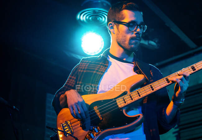 Serious young man playing bass guitar while performing in light club with neon illumination — Stock Photo