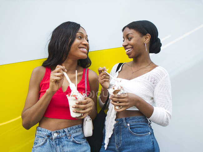 Amigas afro-americanas felizes com copos de sorvete doce olhando umas para as outras enquanto desfrutam de deliciosas sobremesas perto da parede — Fotografia de Stock