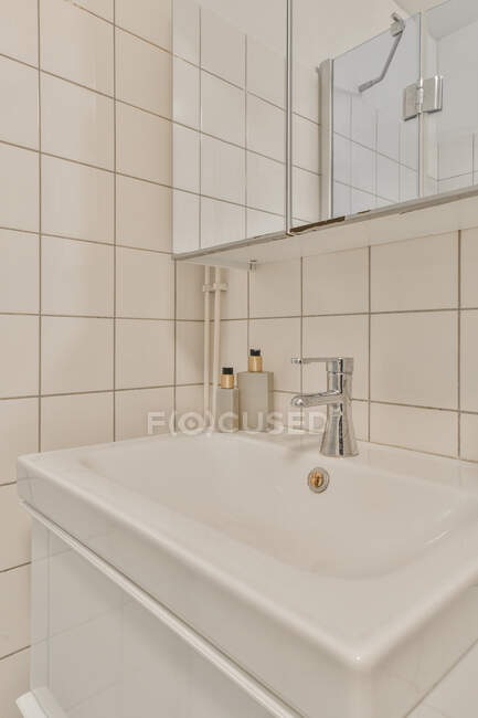 Керамическая раковина с краном и принадлежности помещены на стену с зеркалом в светлом просторном туалете с плиткой стены в квартире — стоковое фото