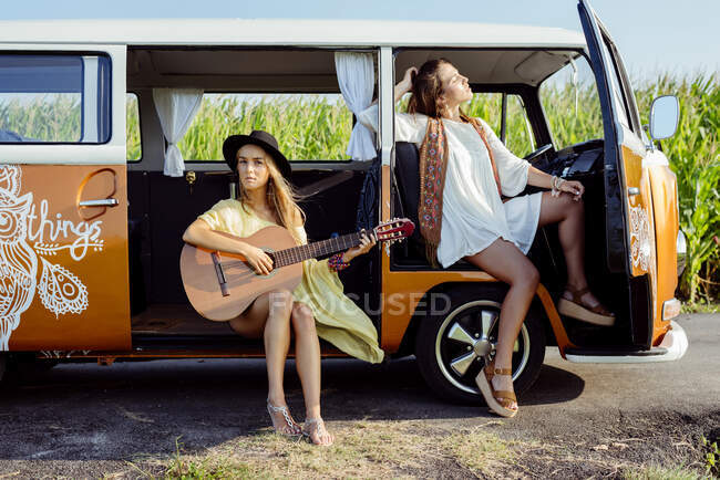 Two pretty girls dressed in summer clothes having fun outside a van and playing guitar — Stock Photo