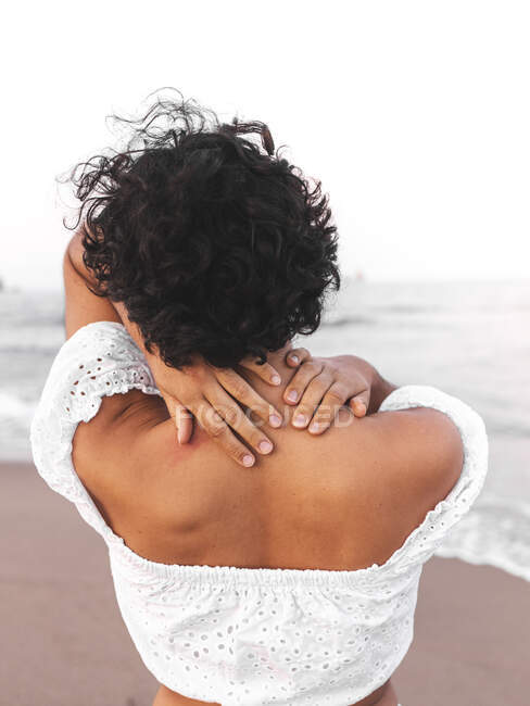 Back view of ethnic female in white stylish outfit and with curly hair standing on seashore and laughing — Stock Photo