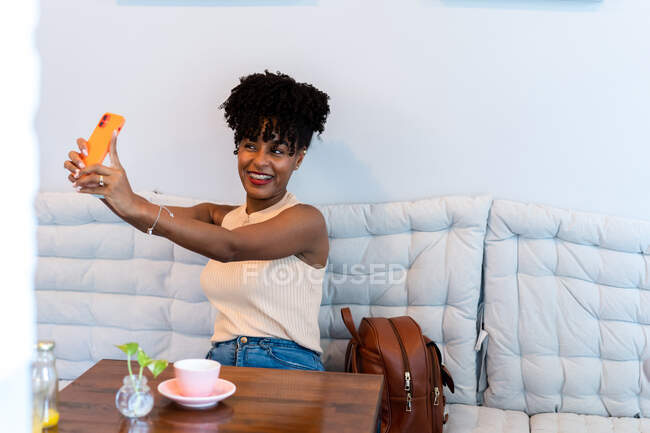 Позитивная молодая афроамериканка с темными вьющимися волосами в повседневной одежде улыбается, делая селфи на смартфоне, сидя за столом с чашкой кофе в современном светлом кафе — стоковое фото