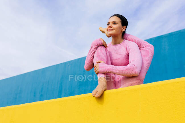 Молодая задумчивая женщина делает вариации Baby Cradle позы во время медитации в йоге asana на синем и желтом фоне — стоковое фото