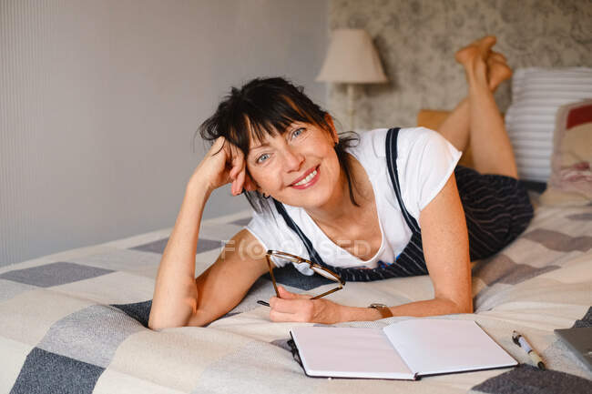 Femme d'âge moyen positive penchée sur la main alors qu'elle était couchée sur le lit avec un bloc-notes et des lunettes ouverts et regardant la caméra — Photo de stock