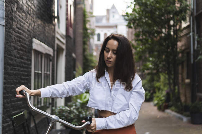 Молодая женщина в повседневной одежде стоит с велосипедом рядом со зданием на улице в центре города, глядя вниз — стоковое фото