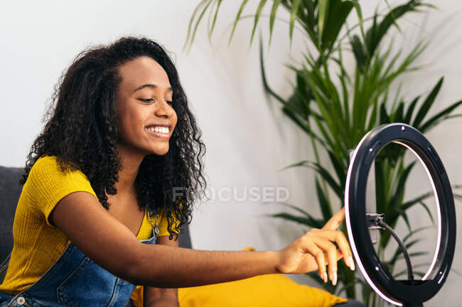 African American woman in denim clothes smiling while sitting on sofa and touching smartphone on glowing ring lamp — Stock Photo