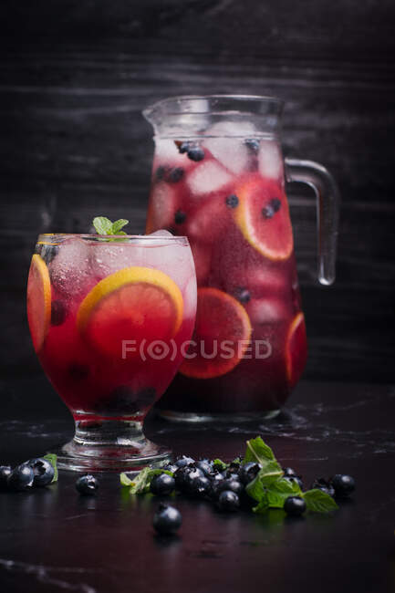 Bicchiere e brocca con rinfrescante limonata fredda con mirtilli freschi e fette di limone poste su tavolo scuro — Foto stock