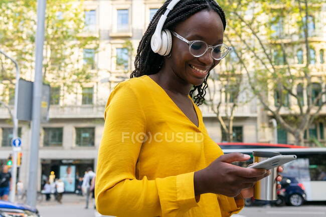 Delighted African American female with thermo mug listening to music in headphones while text messaging on cellphone on street in city — Stock Photo