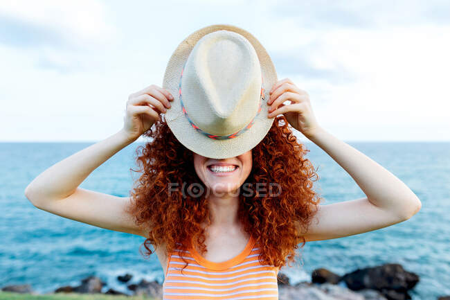 Неузнаваемая женщина с длинными рыжими кудрями, покрывающими лицо шляпой на берегу синего моря — стоковое фото