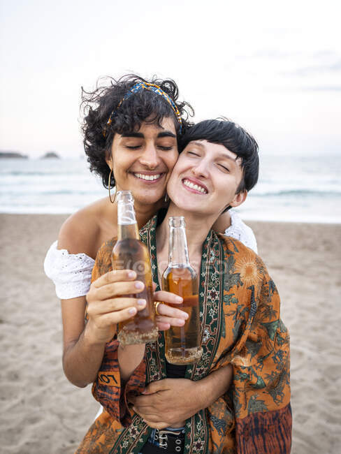 Amando pareja multiétnica de hembras bebiendo cerveza y disfrutando del día de verano en la orilla del mar - foto de stock