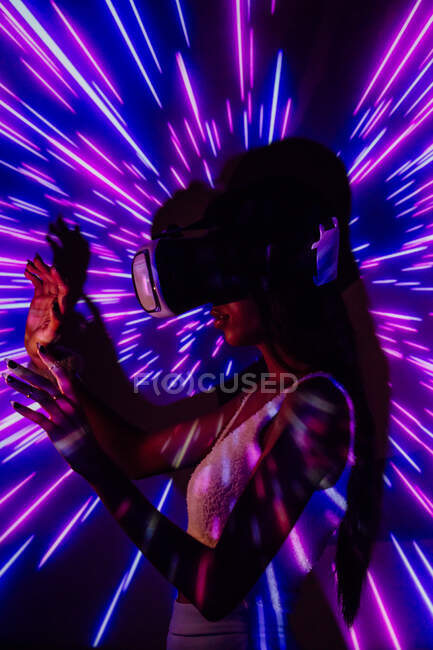 Seitenansicht einer Frau, die im modernen Headset virtuelle Realität erkundet, während sie in rosa und blauen Projektorlichtern steht — Stockfoto