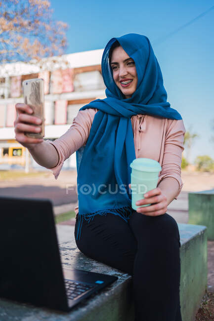 Conteúdo Freelancer árabe feminino em headfast tirar auto-retrato no celular enquanto está sentado no banco com laptop e fazer uma pausa durante o trabalho remoto — Fotografia de Stock
